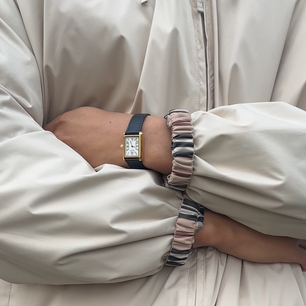 Person wearing a beige jacket with arms crossed with a Gold Monarch Timepiece on, standing against a light blue wooden panel background.